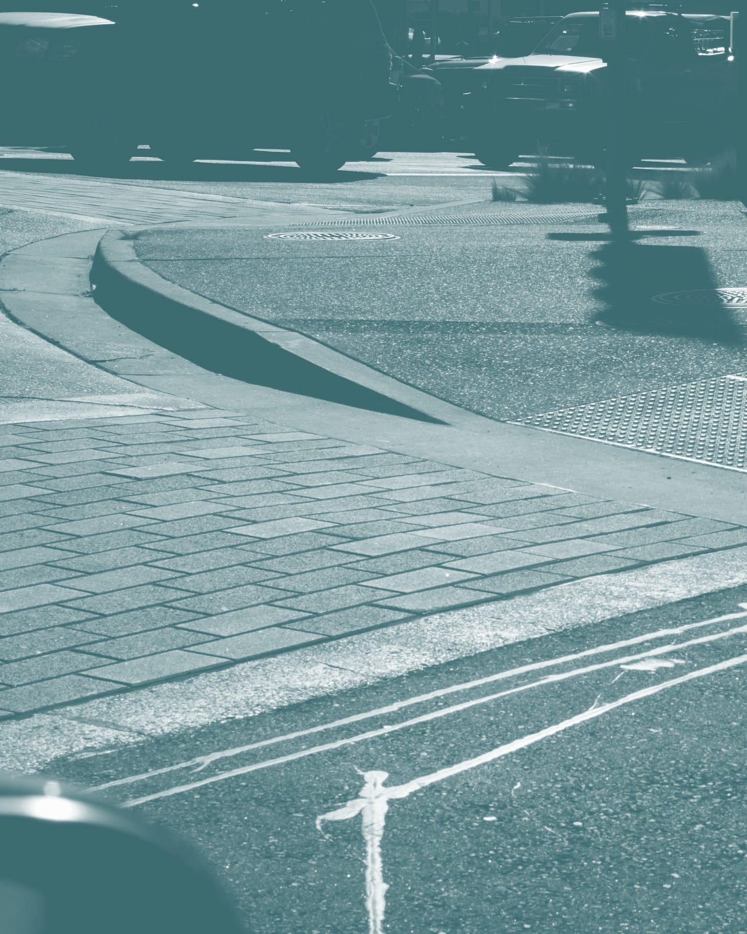 A curb cut ramp at a street corner, sloping from the brick sidewalk down to the crosswalk, with a tactile warning pad on the right and parked vehicles beyond.