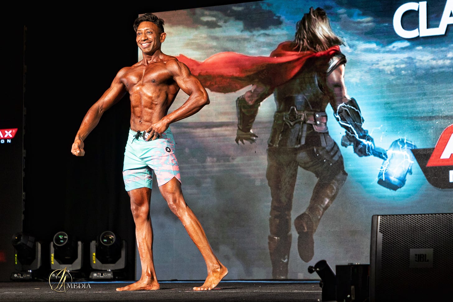 Dr Shiv Goel onstage mid-pose at a physique competition, under bright stage lights.