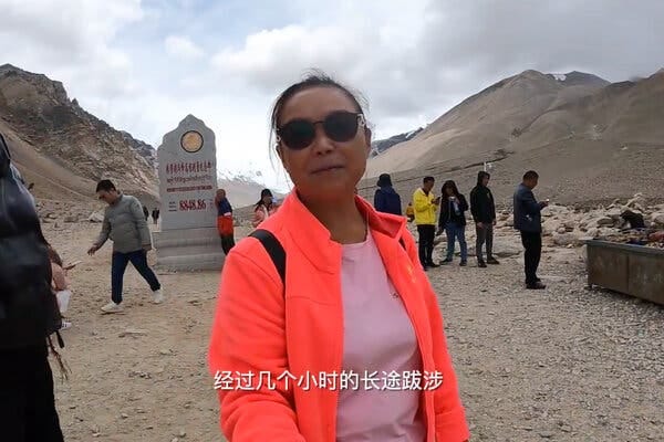 A woman in a bright jacket stands high in the mountains, with a few other people standing behind her. The hills behind her are barren. 