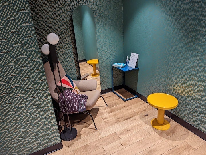 Wellness room with comfortable chair, blanket, pillow, mirror, lamp and two small tables, one of which has sanitary wipes and hand sanitizer