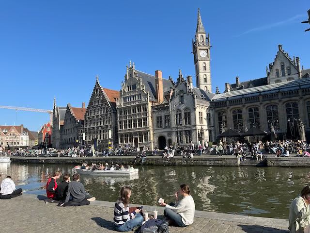 A vibrant scene along the Graslei in Ghent, Belgium, featuring historic guild houses with intricate façades and a prominent clock tower under a clear blue sky. People are sitting along the canal’s edge, chatting and enjoying the sunshine, while others are boating on the water. The lively atmosphere is enhanced by outdoor cafes and groups of people gathered along the quay.