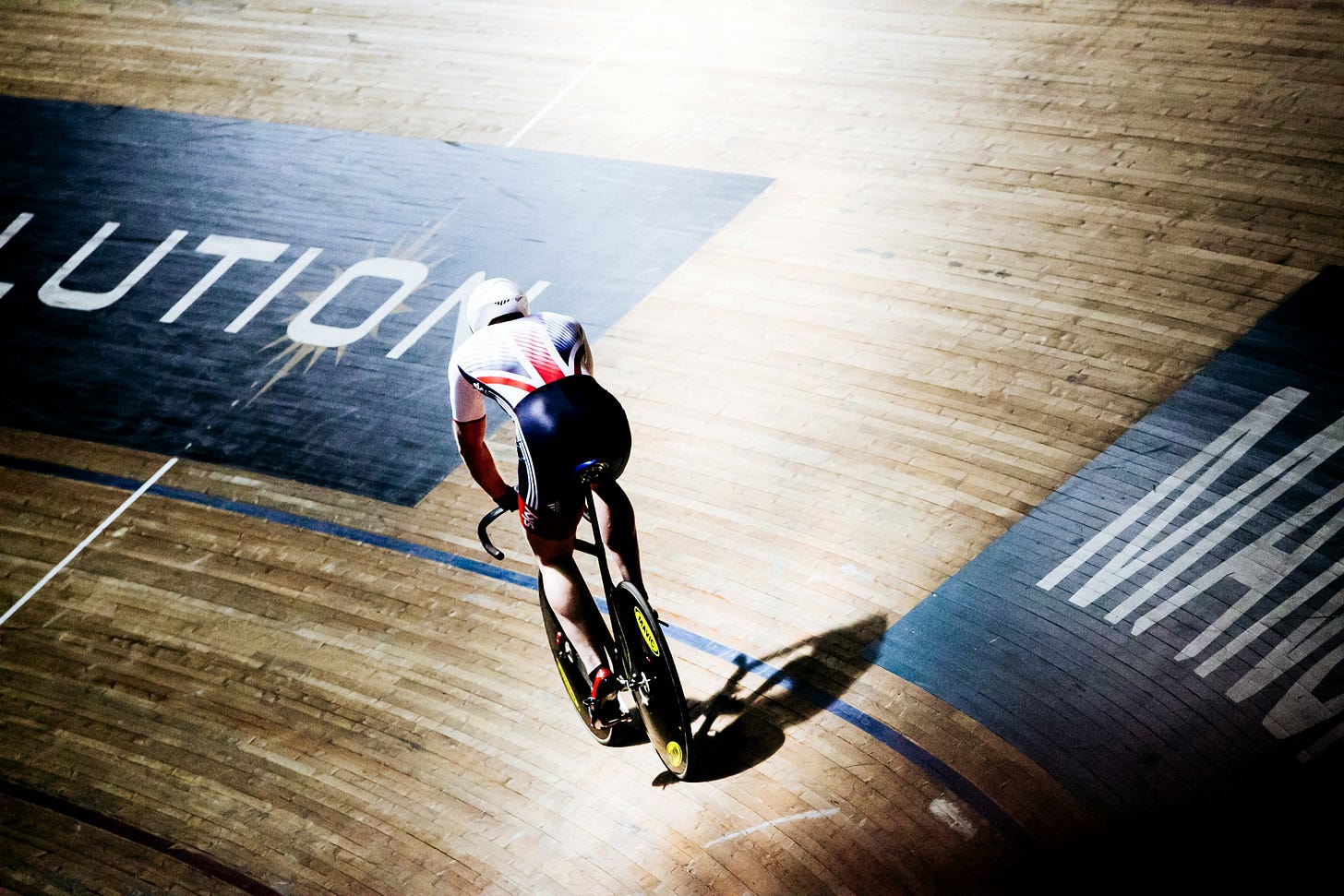 A cyclist rides on a wooden track in a velodrome. We are looking at the rider's back.