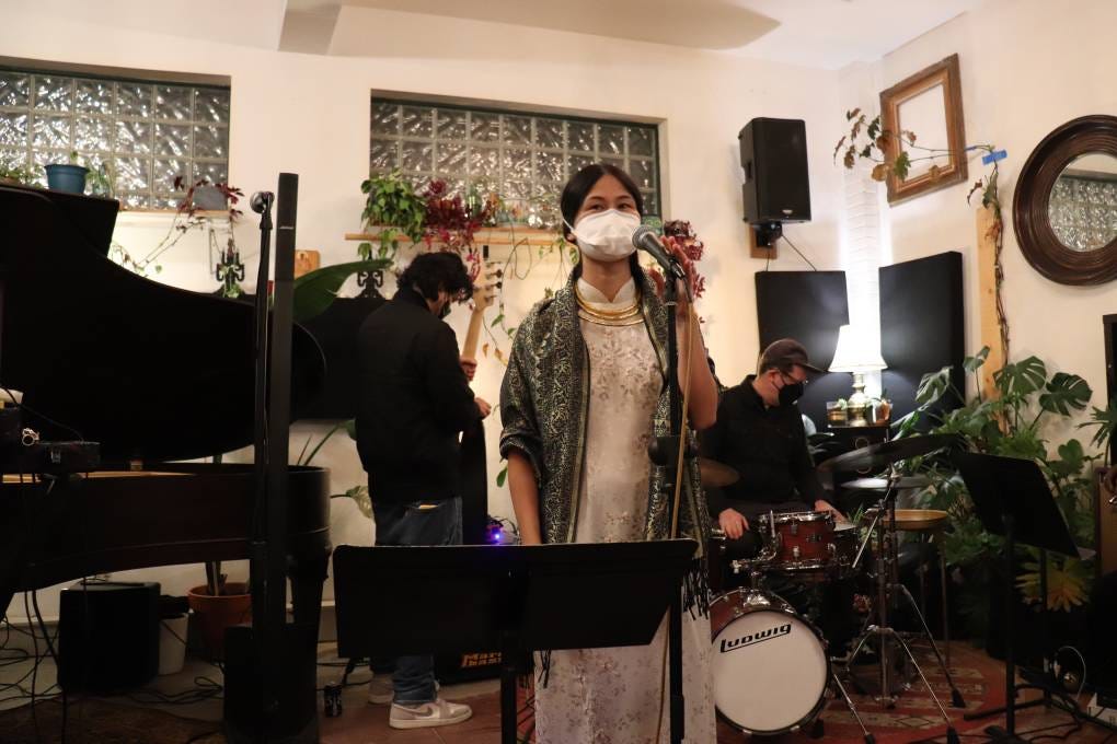 Photo de l'artiste Camille Mai devant un micro sur pied et portant un masque bec de canard blanc, dans une salle avec des plantes, devant des musiciens à la batterie et la guitare masqués aux aussi
