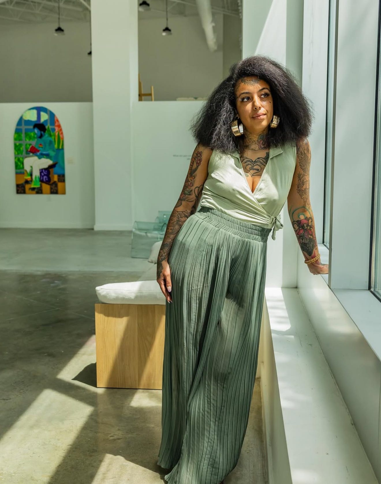 Honey Pierre standing by a sunlit gallery window with one of her blue-figure works in the background.