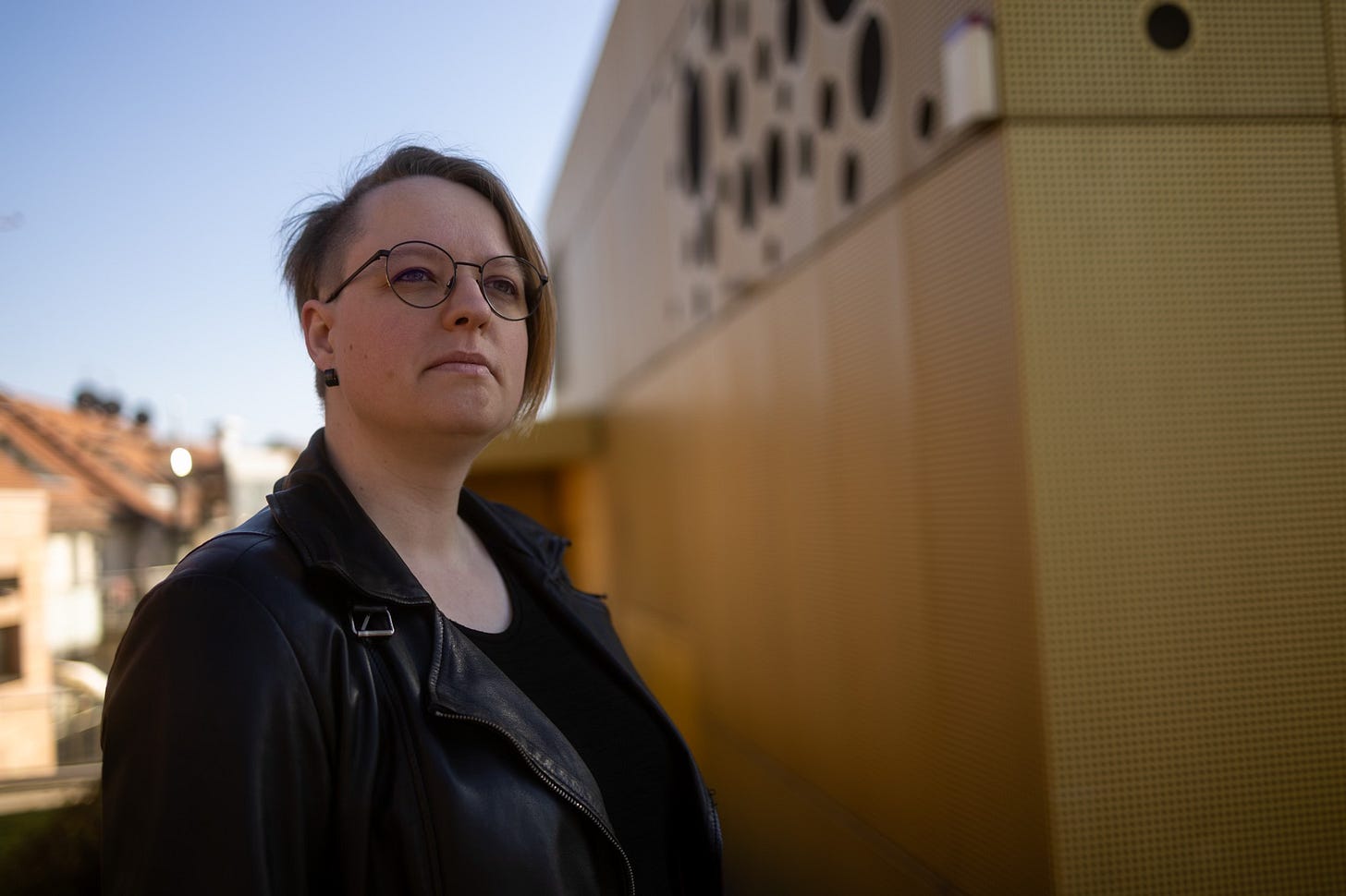 Sibylle steht links im Bild und schaut nach oben. Sie trägt eine schwarze Lederjacke. Links hinter ihr sind Häuser und ein blauer Himmel. Rechts ist ein goldenes Gebäude mit gepunkteter Fassade.