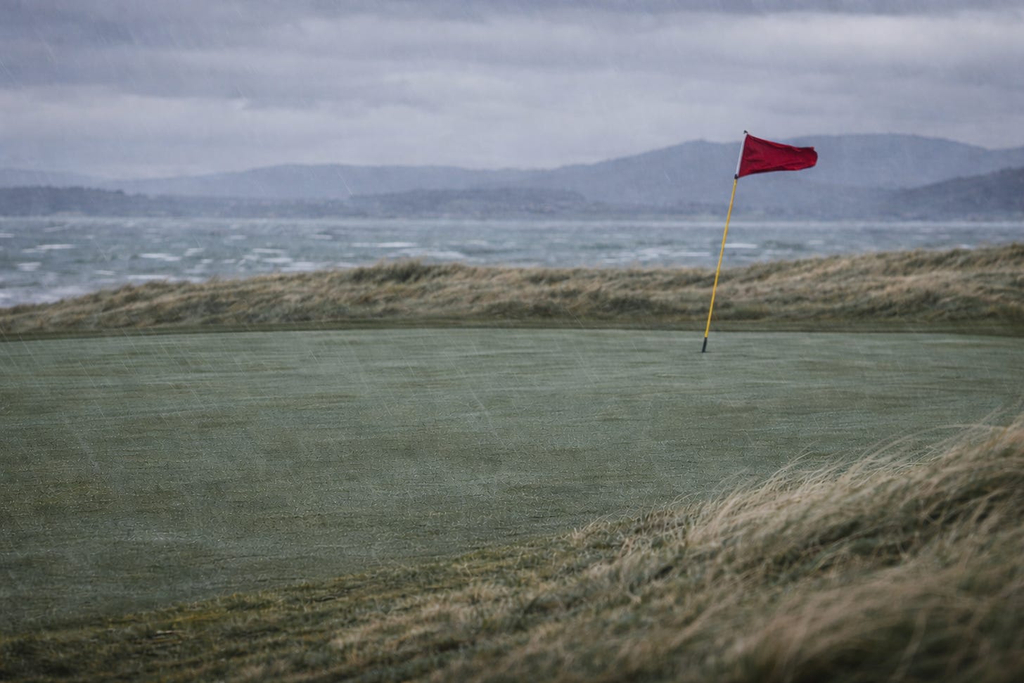 A windswept links green in the rain, with the flag bending in the wind.