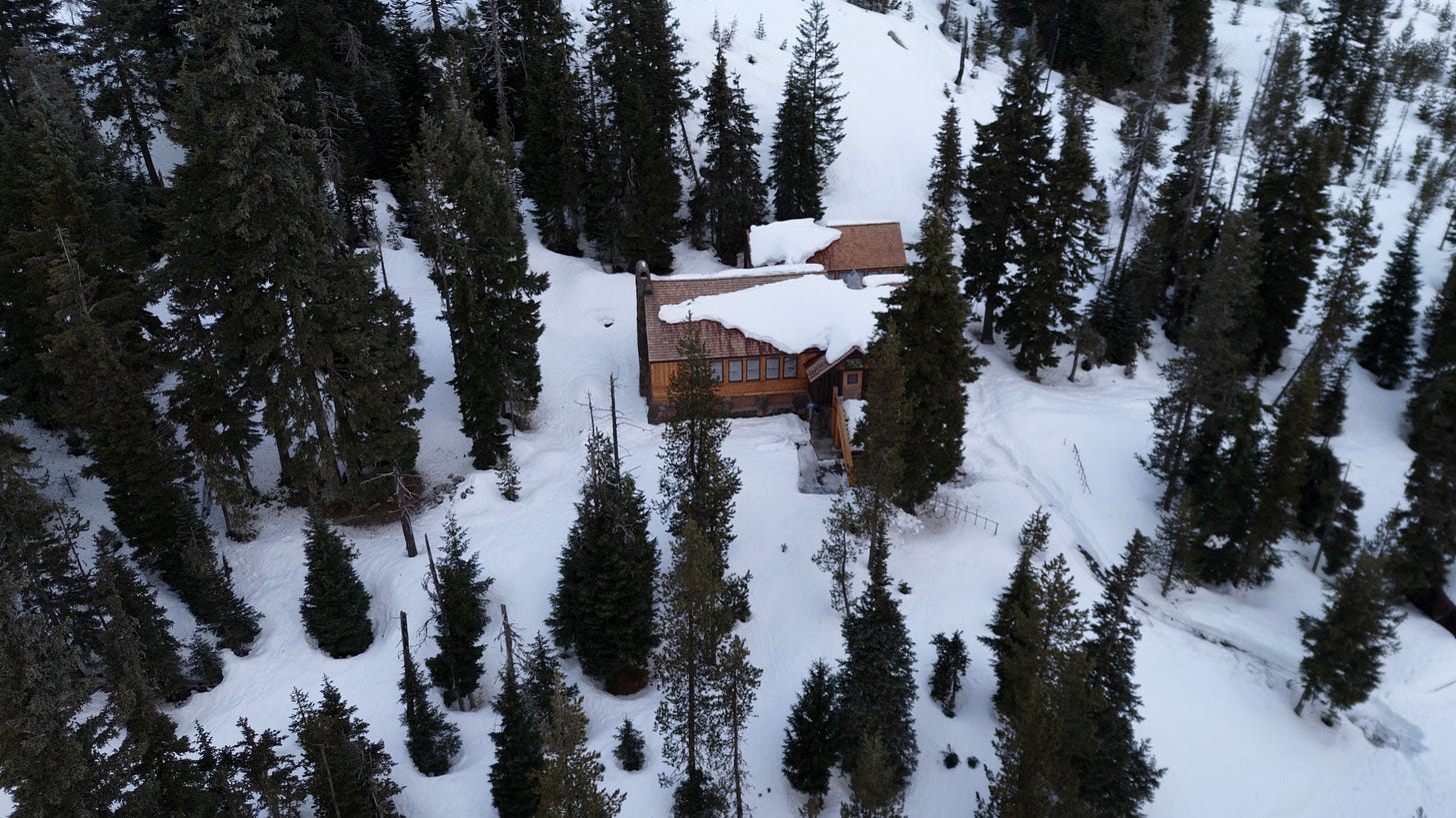 Aerial view of a wooden mountain lodge surrounded by tall evergreen trees and deep snow. The cabin’s roof is partially covered with snow, and narrow paths are visible leading through the snow-covered clearing. The surrounding forest and gentle slopes create a peaceful winter landscape.