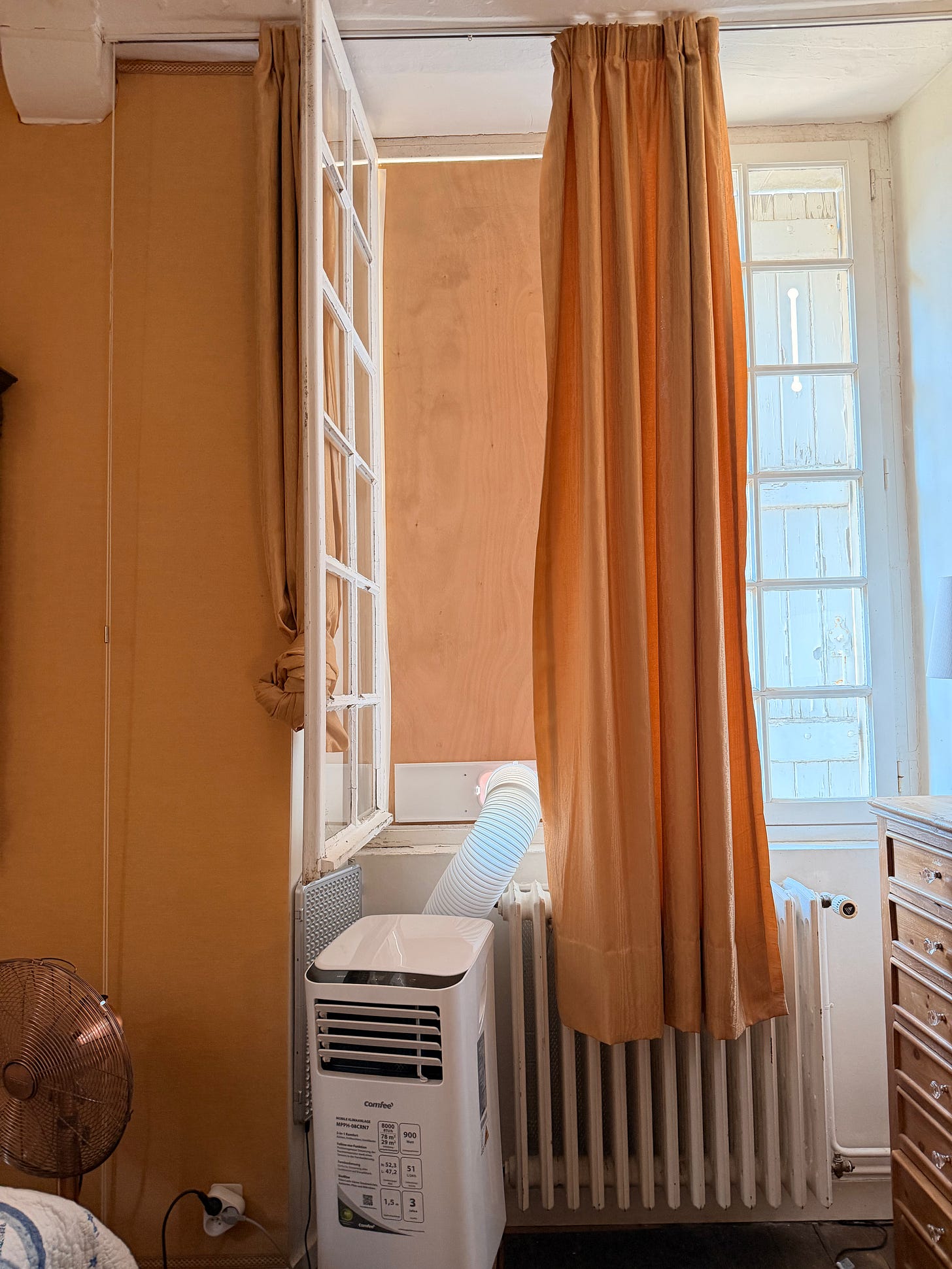A white portable air conditioning unit on a bedroom floor, its exhaust hose running to a tall single-pane window fitted with a custom-cut wooden panel, next to an old radiator and orange curtains. A white portable air conditioning unit on a bedroom floor, its exhaust hose running to a tall single-pane window fitted with a custom-cut wooden panel, next to an old radiator and orange curtains.