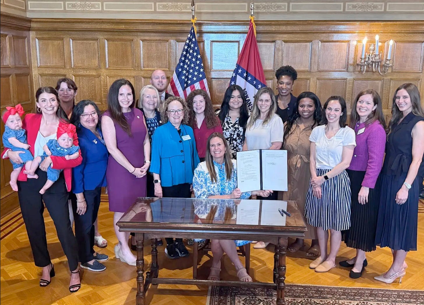 Governor Sanders sits at a glass top desk holding up the bill she signed, while a group of 14 women, one man, and two babies stand behind her smiling in a wood paneled room. Governor Sanders sits at a glass top desk holding up the bill she signed, while a group of 14 women, one man, and two babies stand behind her smiling in a wood paneled room.