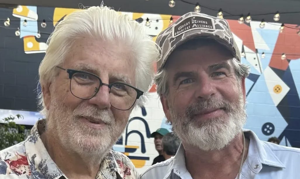 The image shows a man with gray hair wearing a baseball cap and a striped shirt, and another man with a white shirt, both standing near a colorful, geometric patterned wall.

AI-generated content may be incorrect.