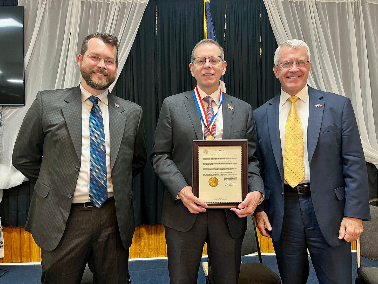 The image shows three men in business attire standing together and smiling at the camera. The man in the center is holding a framed certificate or plaque and is wearing a medal around his neck, suggesting he is being honored or has received an award. There are American flag pins on their suit lapels. The background consists of black and white draped curtains, and there appears to be a United States flag partially visible behind them. No specific recognizable public figures, landmarks, or brands are immediately visible in this image. The image shows three men in business attire standing together and smiling at the camera. The man in the center is holding a framed certificate or plaque and is wearing a medal around his neck, suggesting he is being honored or has received an award. There are American flag pins on their suit lapels. The background consists of black and white draped curtains, and there appears to be a United States flag partially visible behind them. No specific recognizable public figures, landmarks, or brands are immediately visible in this image.