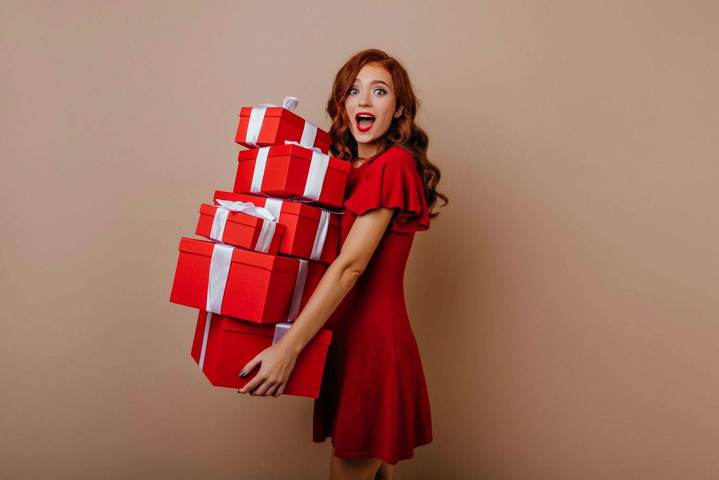 A surprised woman in a red dress holds a tall stack of red gift boxes with white ribbons against a beige background, capturing the excitement and expense of holiday giving. Photo by Look Studio on Unsplash