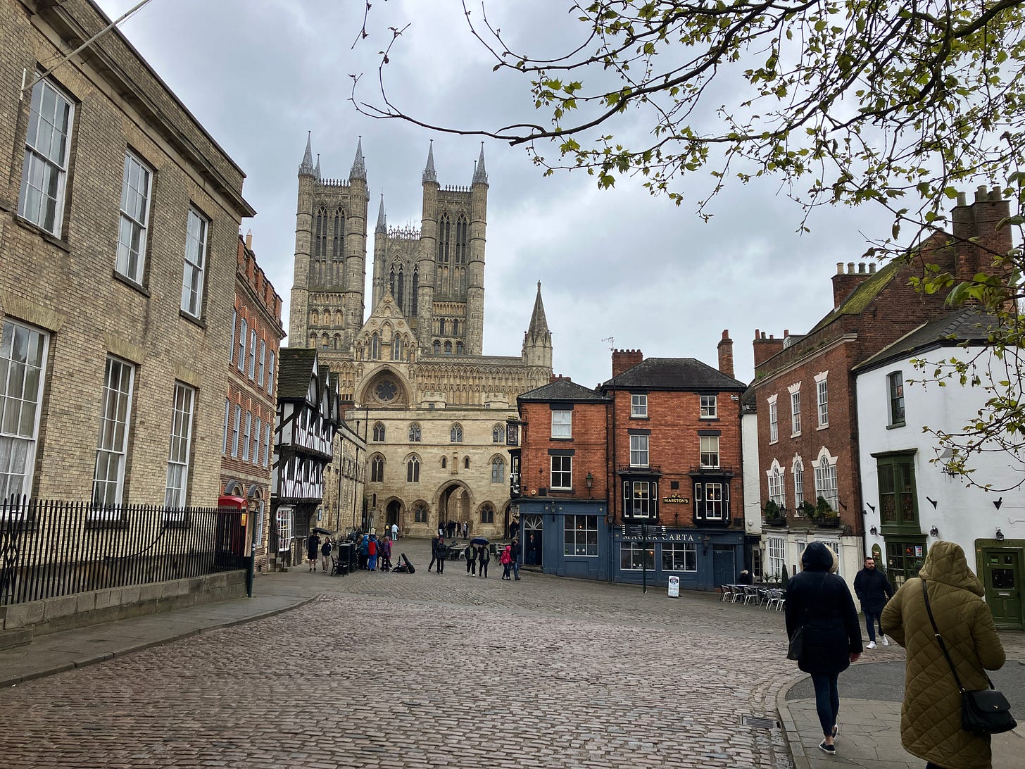Lincoln Cathedral towers over surrounding historic buildings, with people walking on a cobbled street on an overcast day. Lincoln Cathedral towers over surrounding historic buildings, with people walking on a cobbled street on an overcast day.