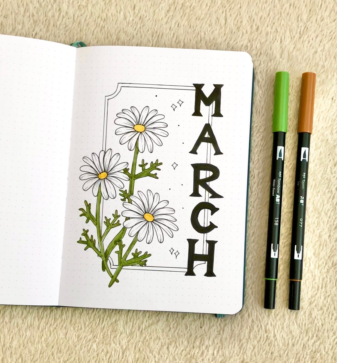 A journal page with “March” written in serif letters and daisy illustrations. Page is next to a brown marker and a green marker.