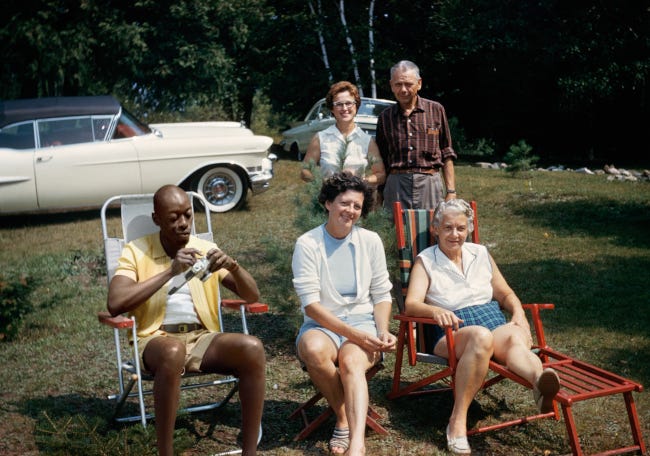 Un groupe d'amis de soixante-soixante-dix ans en camping, assis sur des chaises pliantes pour les uns, debout près d'une grosse voiture américaine pour d'autres. Parmi eux, en short jaune et t-shirt blanc, un jeune homme noir profite lui aussi du soleil.