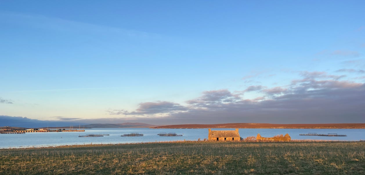A photo of the sea, there is a field and a falling down stone building in the foreground, then the sea and then further land and hills in the distance. It is a bright day with blue sky