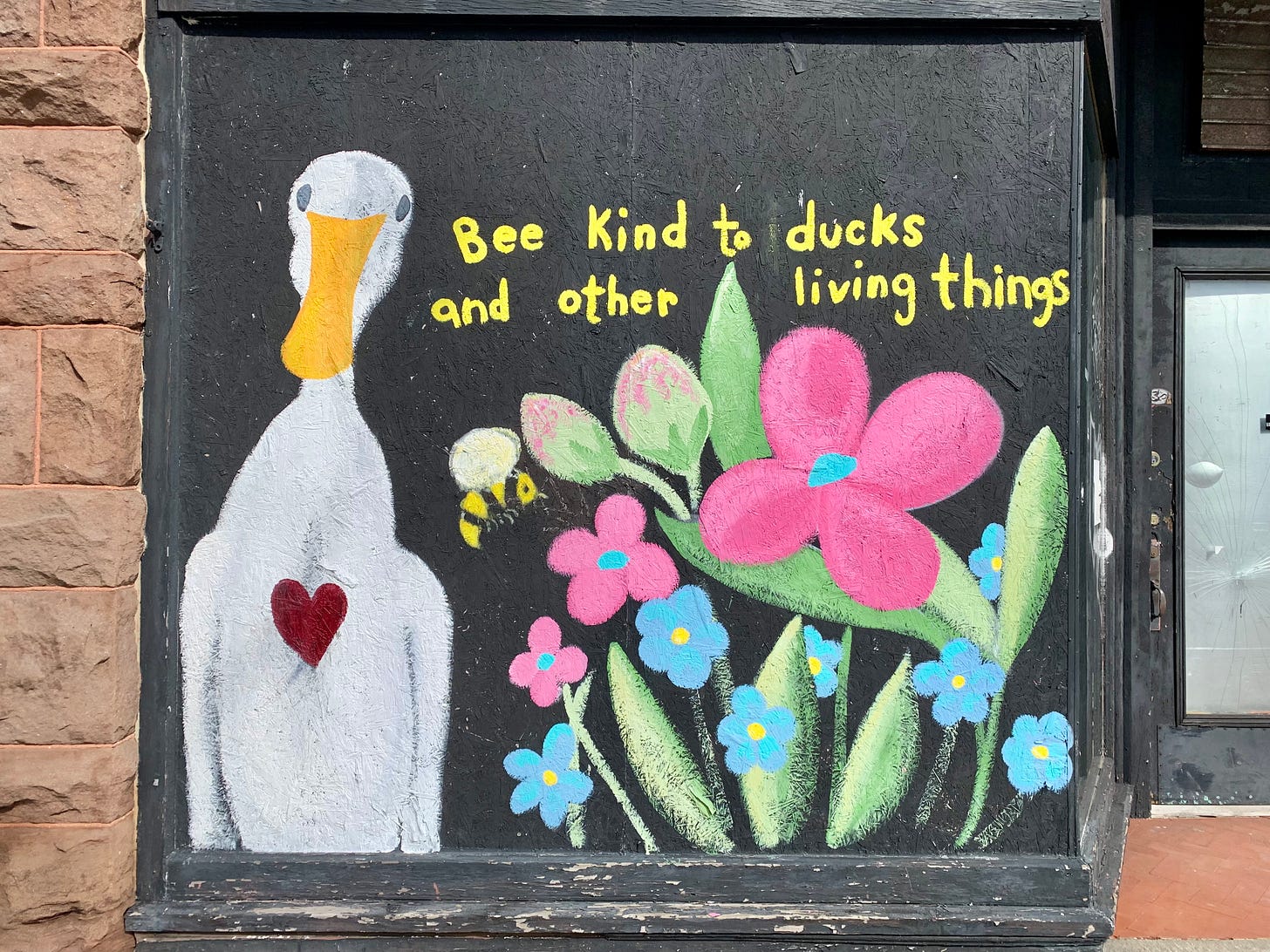 A mural on the side of an abandoned building featuring a duck, flowers, a bee, and the words BEE KIND TO DUCKS AND OTHER LIVING THINGS
