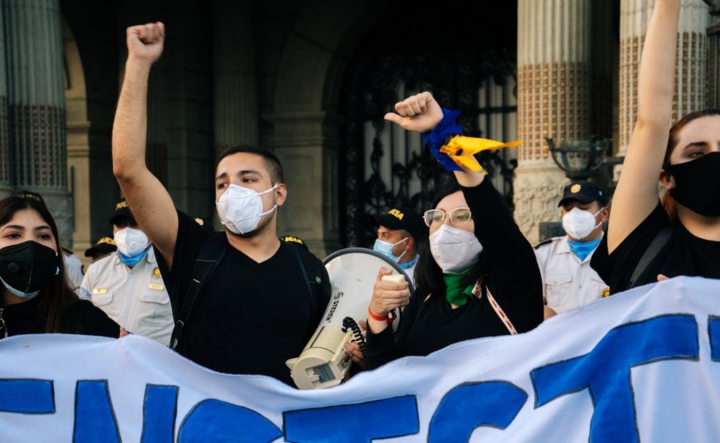 man in black vest wearing white mask holding white and blue flag man in black vest wearing white mask holding white and blue flag