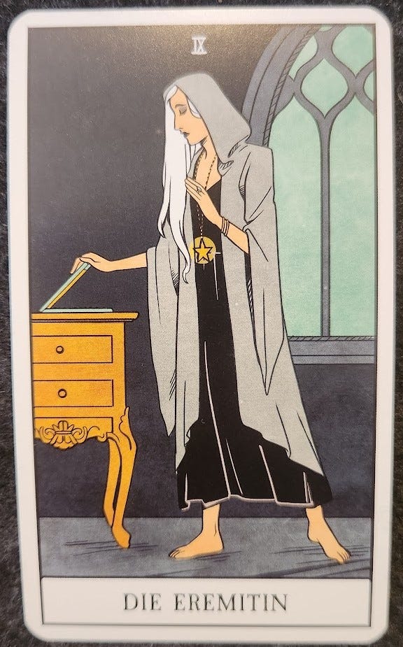 A tarot card labelled IX Die Eremitin. A woman with long white hair wearing a black dress and a long hooded grey cloak is standing in a grey room. She is closing the lid of a laptop, her eyes slightly downcast. She is wearing a long necklace with a pentacle. There is a big glass window behind them, showing a light sky outside.  