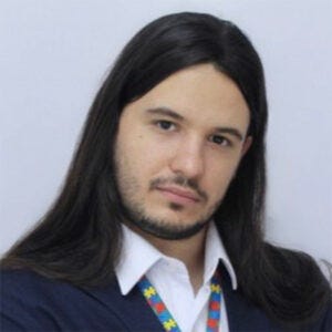 Homem branco, de cabelo castanho escuro comprido, usa terno com um colar de identificação de pessoa com transtorno de espectro autista (TEA). Inclina a cabeça, olhando com expressão séria. Fundo branco.