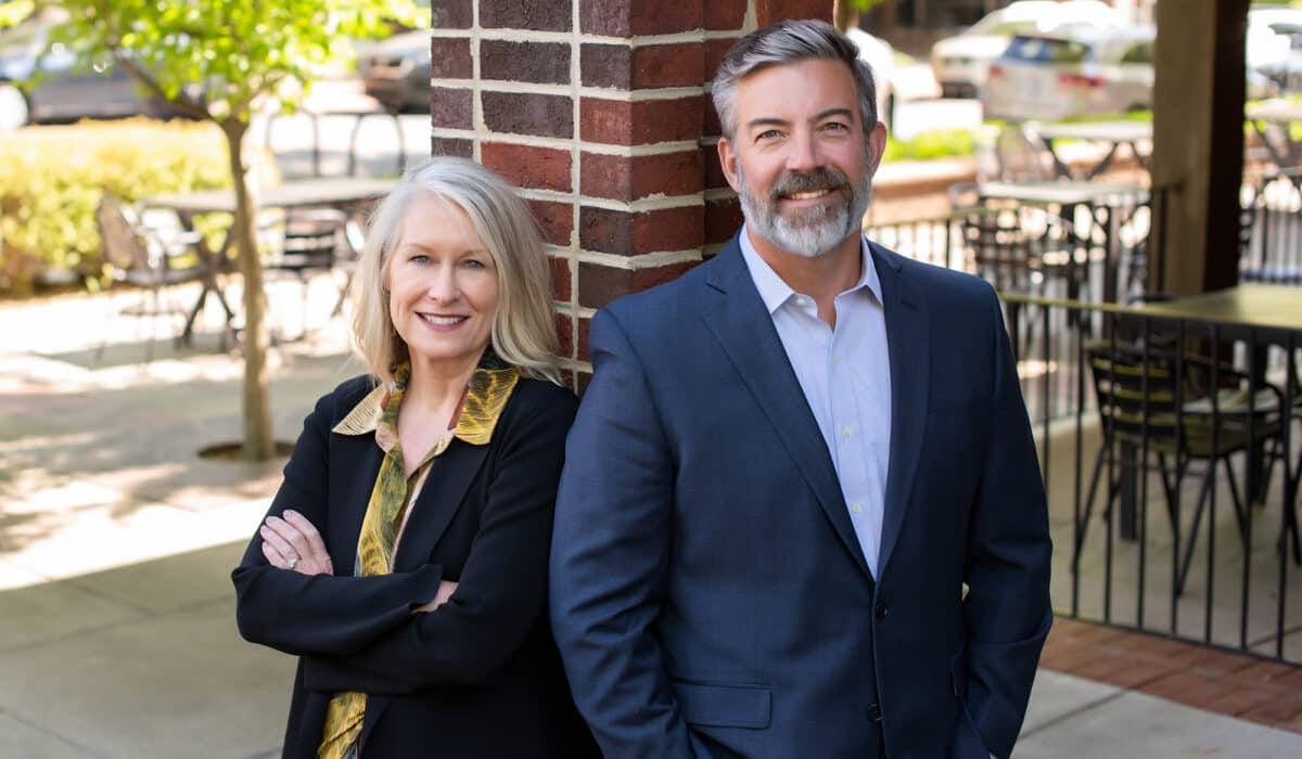 A professional man and woman in business attire stand confidently with their arms crossed, the woman in a black jacket with a yellow accent and the man in a blue suit, outside near a brick column and outdoor seating area.