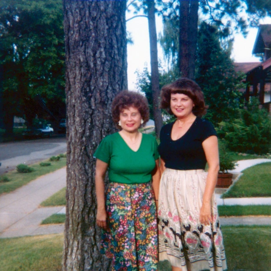 Two women stand beside a large tree, smiling for the camera. They are dressed in colorful skirts and tops, with a residential street and greenery in the background.