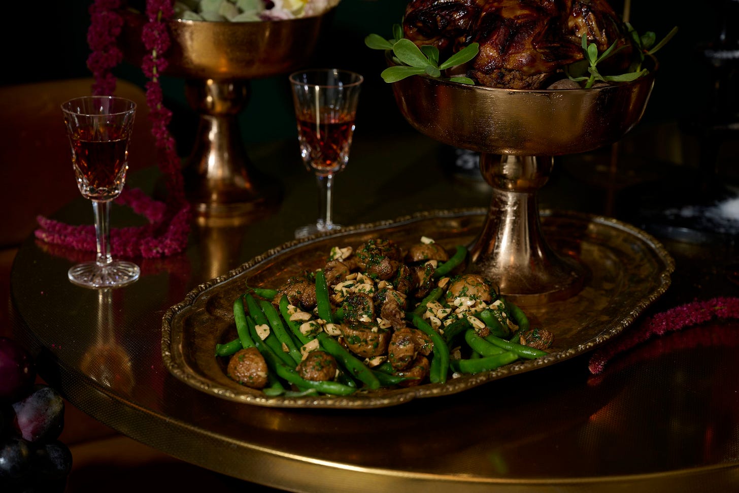 Haricot vert with chanterelle mushrooms and tarragon vinaigrette served on a vintage brass platter, with glasses of rosé vermouth and lacquered quails in the background on a gold pedestal bowl.