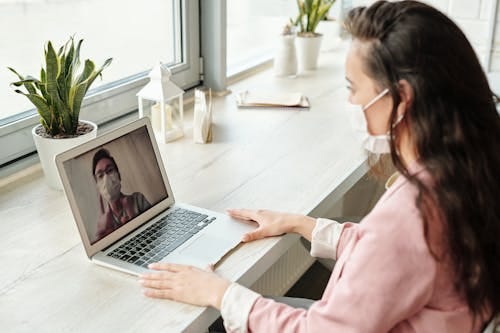 Free Woman and doctor video calling on laptop, both wearing masks, symbolizing telehealth during COVID-19. Stock Photo