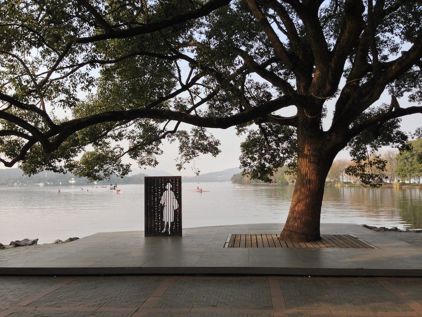 Fotografia em cores de parque à beira de um rio. Sob uma árvore, há uma instalação artística representando, em branco sobre material preto, um perfil feminino. Fotografia em cores de parque à beira de um rio. Sob uma árvore, há uma instalação artística representando, em branco sobre material preto, um perfil feminino.
