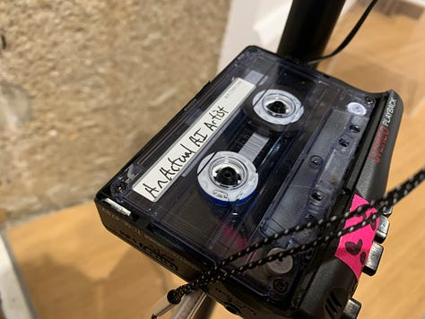 A variety of new media art works using screens, projections, including a large yellow screen and an anaologe cassete with the caption "an actual AI artist" 