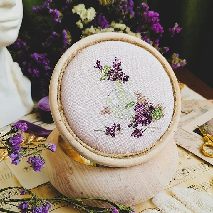 A photograph of a dainty little cross-stitch on a table with flowers that match the cross-stitch piece. A photograph of a dainty little cross-stitch on a table with flowers that match the cross-stitch piece.