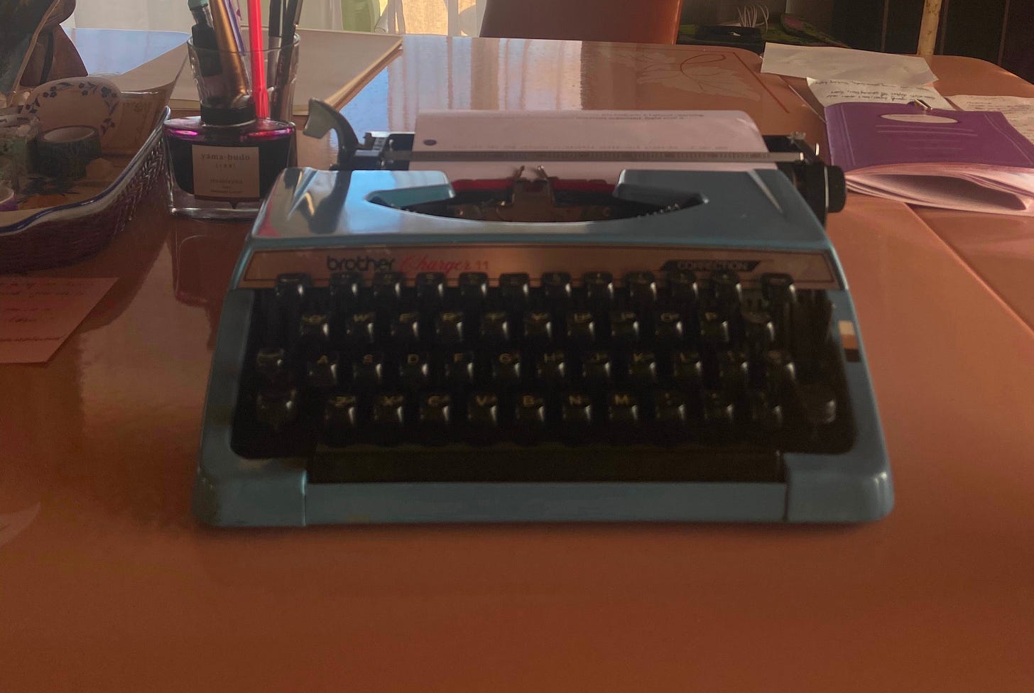Blue brother manual typewriter on a peach colored desk, surrounded by pens and ink. Blue brother manual typewriter on a peach colored desk, surrounded by pens and ink.