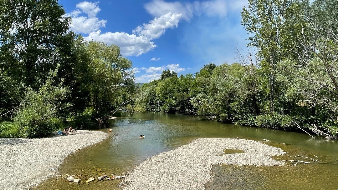 A wide, calm river. In the distance, some people are swimming in the water. In the distance: a blue sky and lush, green trees.
