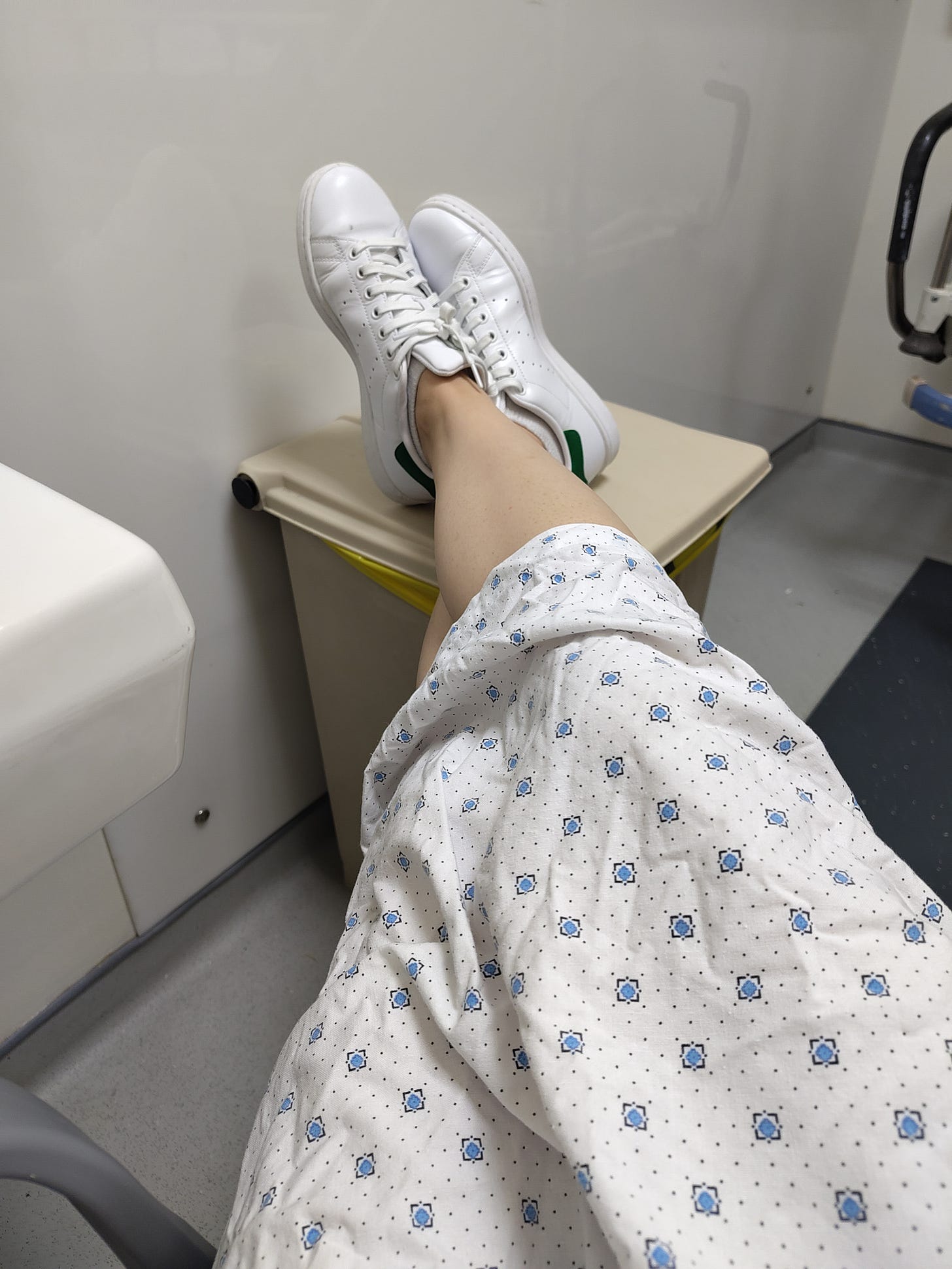 The lower body of a patient in a hospital gown and white tennis shoes in an emergency room. The feet are perched on a garbage bin.