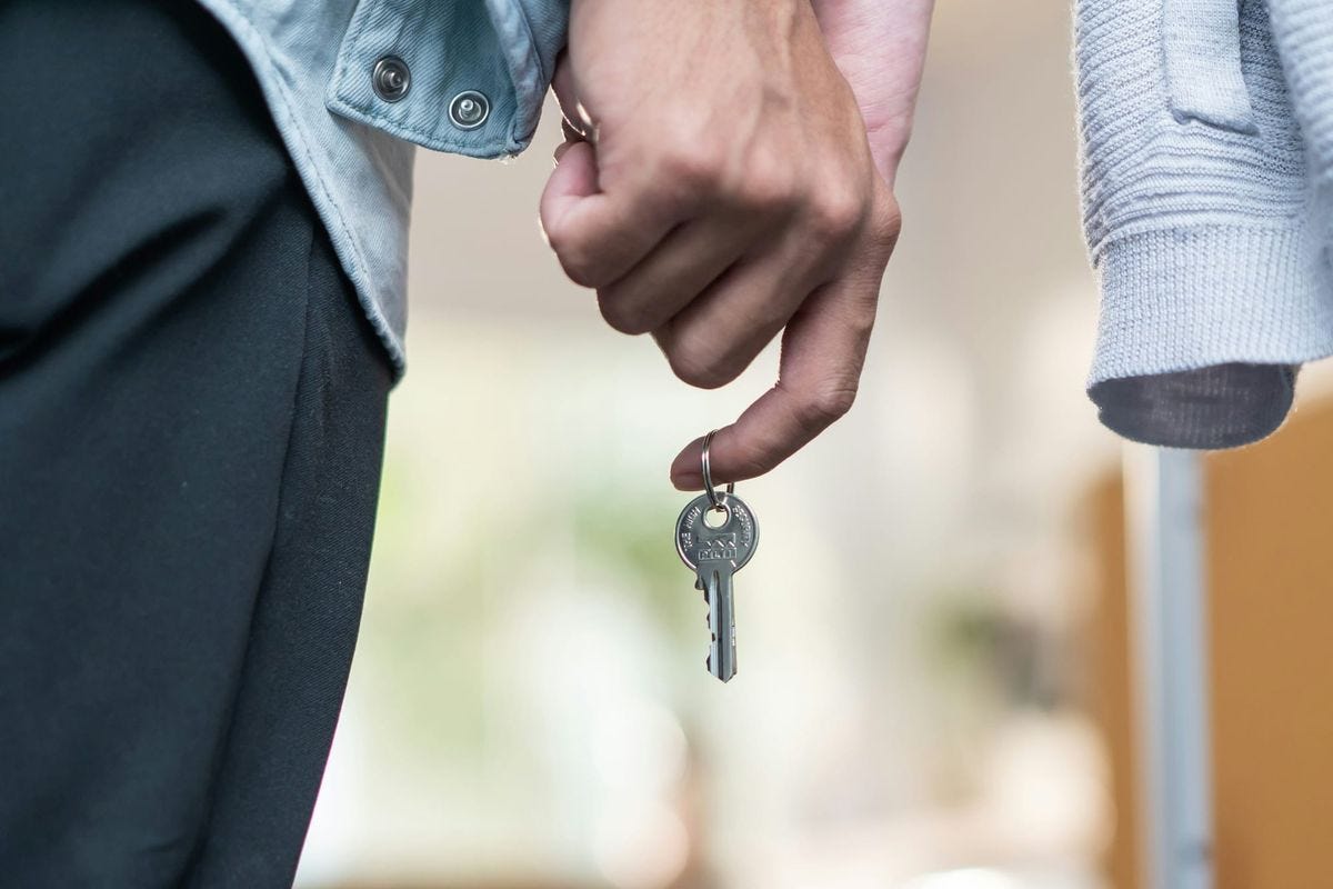 A hand holding a single house key on a ring, with a second person standing alongside. A hand holding a single house key on a ring, with a second person standing alongside.