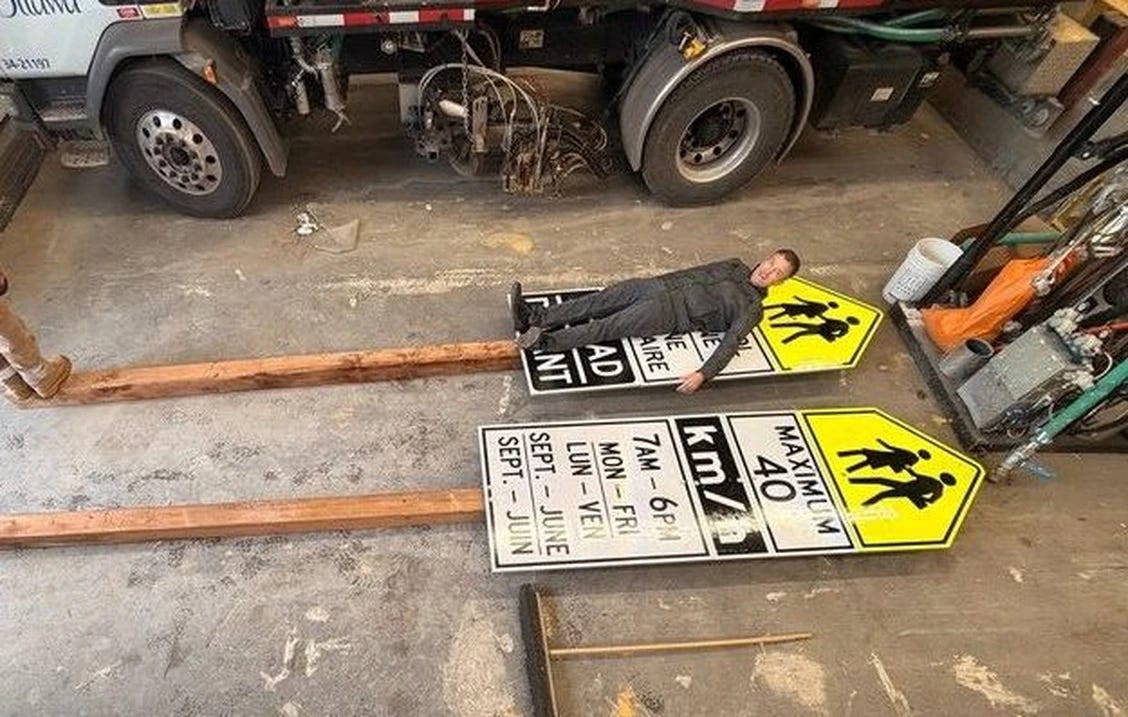 In a garage, a man lays atop a speed limit sign placed on the ground. HIs outstretched body does not cover the whole height of the massive sign. In a garage, a man lays atop a speed limit sign placed on the ground. HIs outstretched body does not cover the whole height of the massive sign.