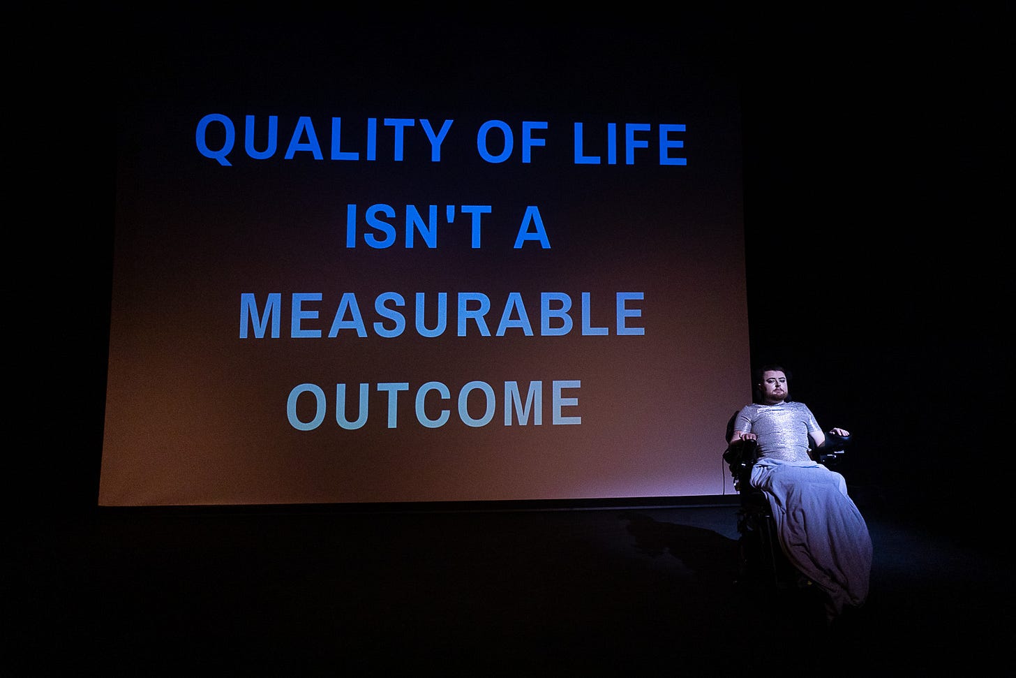 Image: Jamie Hale, a white person with dark red hair and a ginger beard sits in an electric wheelchair at the side of the stage wearing a silver top, with a hospital blanket over their legs. Beside them is projected “Quality of life isn’t a measurable outcome”.