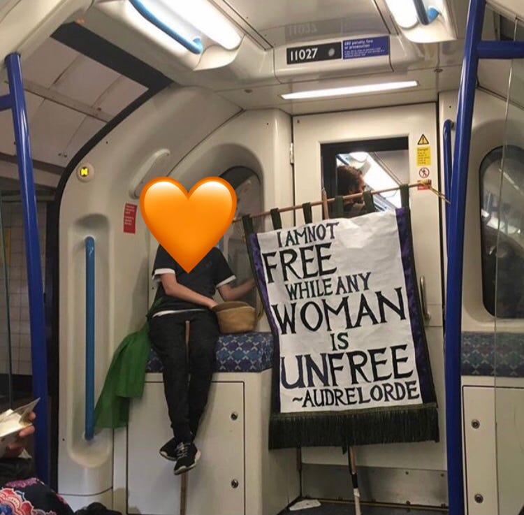 A young teenager holding a sign saying 'I a not free while any women is unfree' - Audre Lorde, whislt sat on the London underground. Face covered with an orange heart. 