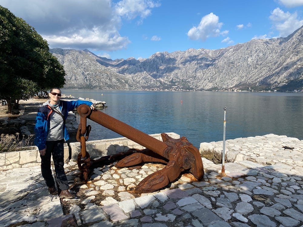 A women stands next to a large rusted anchor on the shore of the water with mountains in the background.