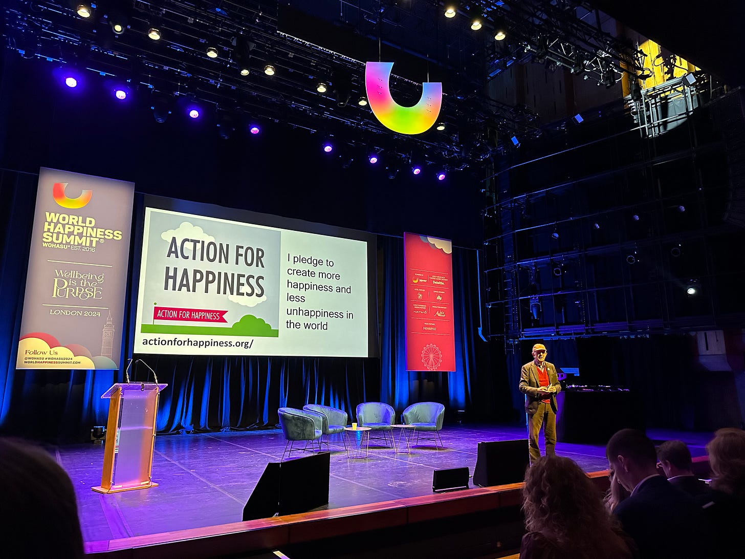 Lord Richard Layard stands on the stage at the WOHASU summit in front of a slide promoting Action for Happiness actionforhappiness.org "I pledge to create more happiness and less unhappiness in the world". 