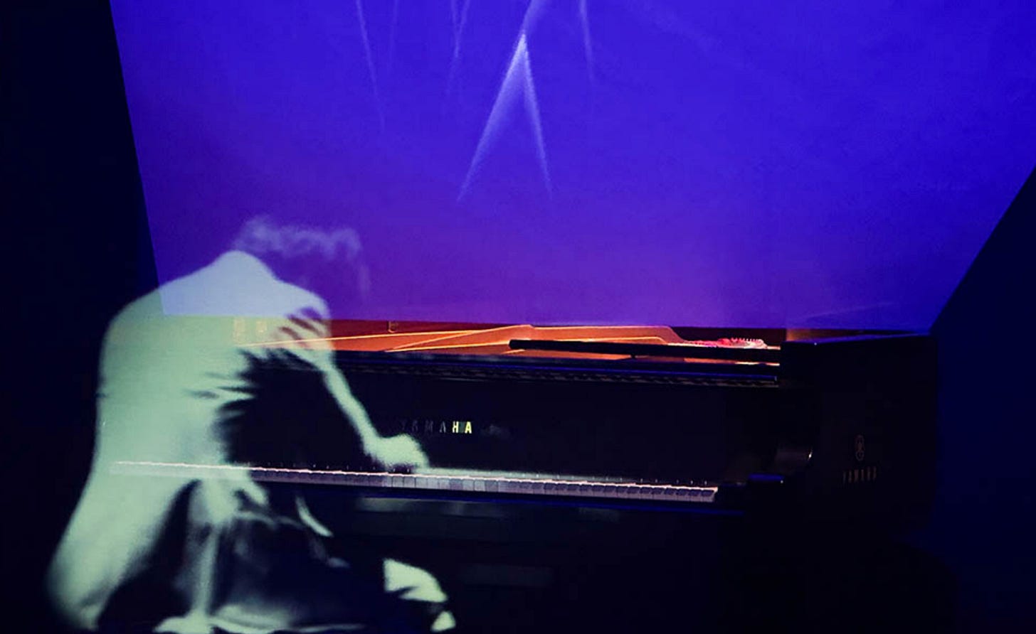 Color photo of a projection of Ryuichi Sakamoto at a player piano. Color photo of a projection of Ryuichi Sakamoto at a player piano.
