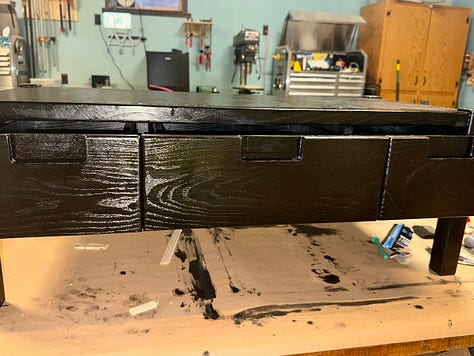 Three views of a wooden bench with drawers. Left: natural wood drawer fronts fitted into the frame. Center: drawers painted dark with visible wood grain. Right: completed bench with smooth black finish across all drawers.