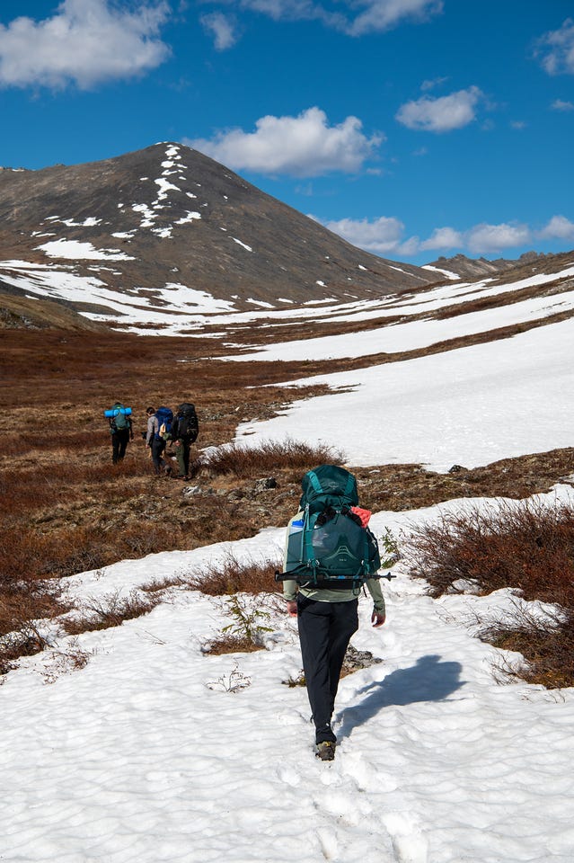 Four hikers with packs move across snow patches and tundra toward a snow-covered slope below a dark peak, one in the foreground crossing a broad snowfield.
