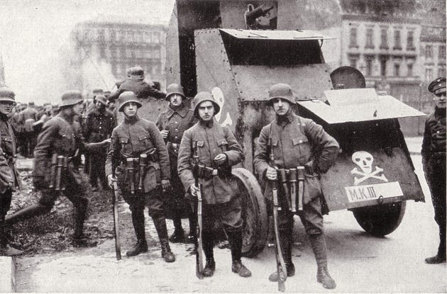 Freikorps troops with weapons pose in front of an armored car with a Totenkopf. Behind them, smoke rises.