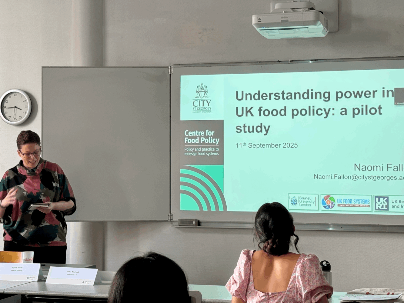 Woman in front of screen showing presentation slide. Text on slide: Understanding power in UK food policy: a pilot study. 