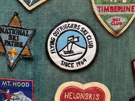 Left: A pinback button about 1" across sits in a museum display case. It was for the Jaycee Ski School and is red, white, and blue. Center: A sit-ski outrigger from the 1990s. It has an aluminum shaft and the very front tip of a ski attached to the end. Right: A white, black, and light blue button for the flying outriggers ski club. Their logo shows a pair of standing-ski outriggers, like crutches, with flags on them, waving in the wind..