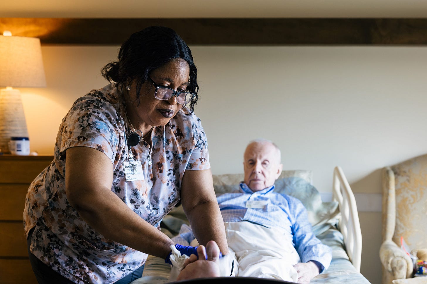 A senior man is lying in a bed in a nursing home. A nurse is carefully helping him with his socks. A senior man is lying in a bed in a nursing home. A nurse is carefully helping him with his socks.