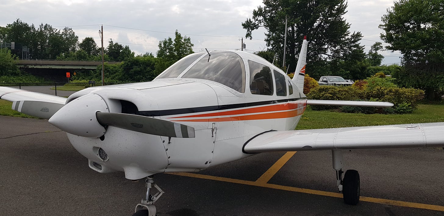 A small single-engine plane parked on an airfield apron, with a runway and trees behind it. A small single-engine plane parked on an airfield apron, with a runway and trees behind it.