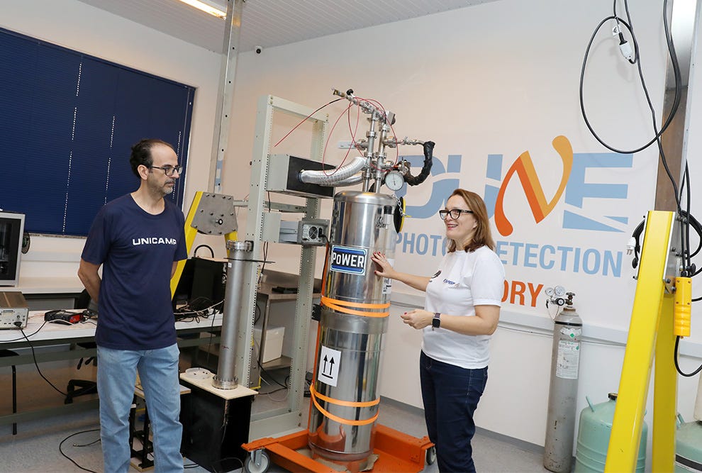Duas pessoas em um laboratório observam um equipamento cilíndrico metálico prateado com fitas laranja, posicionado sobre uma base laranja, sendo que a pessoa à esquerda veste camiseta azul-marinho com texto "UNICAMS" e a pessoa à direita veste camiseta branca, ambas diante de estruturas metálicas de suporte, cabos, e um painel de parede com logo laranja e texto azul parcialmente visível contendo as palavras "PHOTO DETECTION" e "ORY". Duas pessoas em um laboratório observam um equipamento cilíndrico metálico prateado com fitas laranja, posicionado sobre uma base laranja, sendo que a pessoa à esquerda veste camiseta azul-marinho com texto "UNICAMS" e a pessoa à direita veste camiseta branca, ambas diante de estruturas metálicas de suporte, cabos, e um painel de parede com logo laranja e texto azul parcialmente visível contendo as palavras "PHOTO DETECTION" e "ORY".