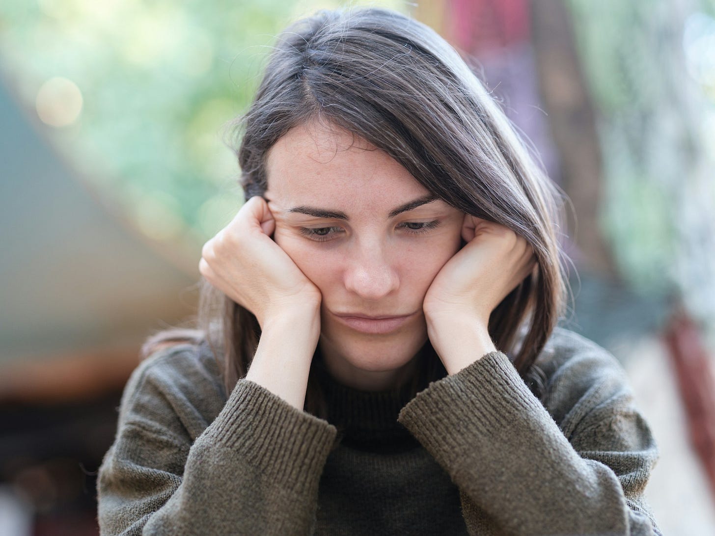 The image shows a person with long dark hair looking downcast with their face resting in their hands. They appear to be in a contemplative or distressed state, wearing a grey jumper in what seems to be an indoor setting with a blurred background. The posture and facial expression suggest feelings of sadness, worry, or exhaustion.